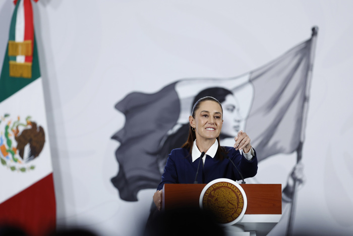 La presidenta Claudia Sheinbaum en conferencia de prensa matutina señalando desde el podio con la bandera de México al fondo.