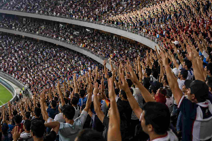 Afición mexicana de pie en las tribunas de un estadio alzando los brazos durante un partido oficial.