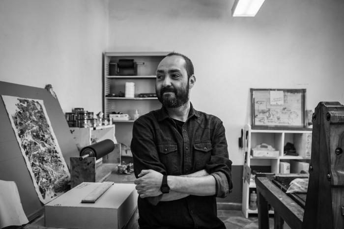 El artista visual y crítico de arte michoacano Raúl Calderón Gordillo posa sonriendo en su taller de estampa y grabado en blanco y negro.