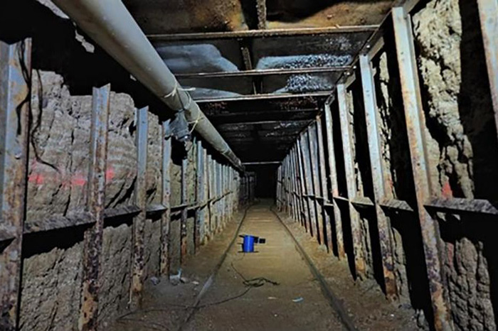 Interior de un túnel subterráneo transfronterizo iluminado, con paredes reforzadas y sistema de rieles en la zona fronteriza.