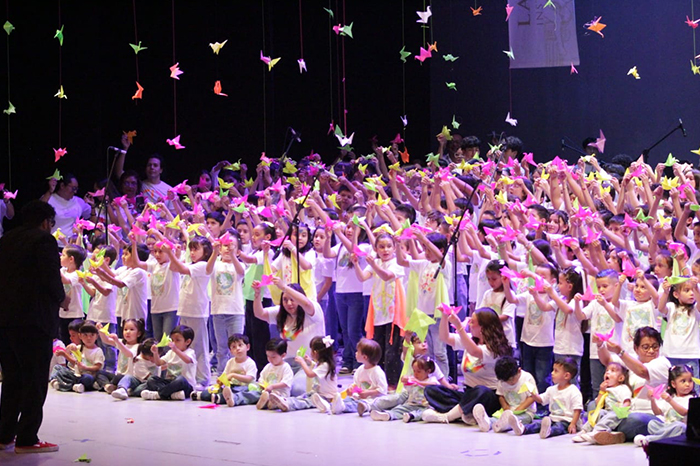 Estudiantes de preescolar y primaria sobre el escenario del Teatro Morelos cantando en coro rodeados de grullas de papel origami.