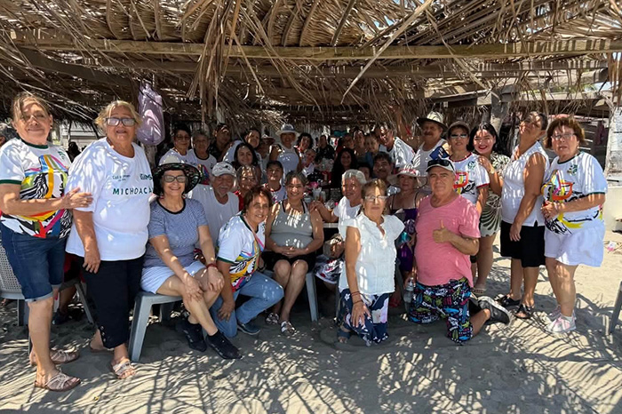 Grupo de adultos mayores conviviendo y sonriendo en un ambiente recreativo frente al mar.