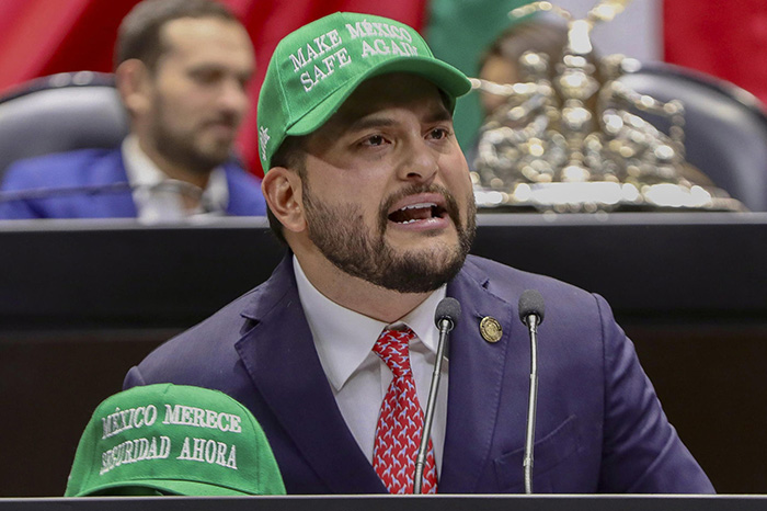 Diputado en la tribuna de la Cámara de Diputados portando una gorra verde con la leyenda Make México Safe Again durante un debate político.