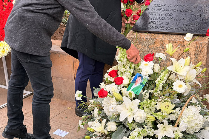 Manos colocando flores y un arreglo floral conmemorativo junto a la placa en honor a las víctimas del atentado del 15S en Morelia.